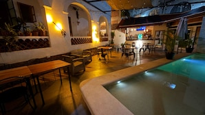 Un área de piscina interior con asientos y mesas, rodeada de paredes con arcos y plantas en macetas.