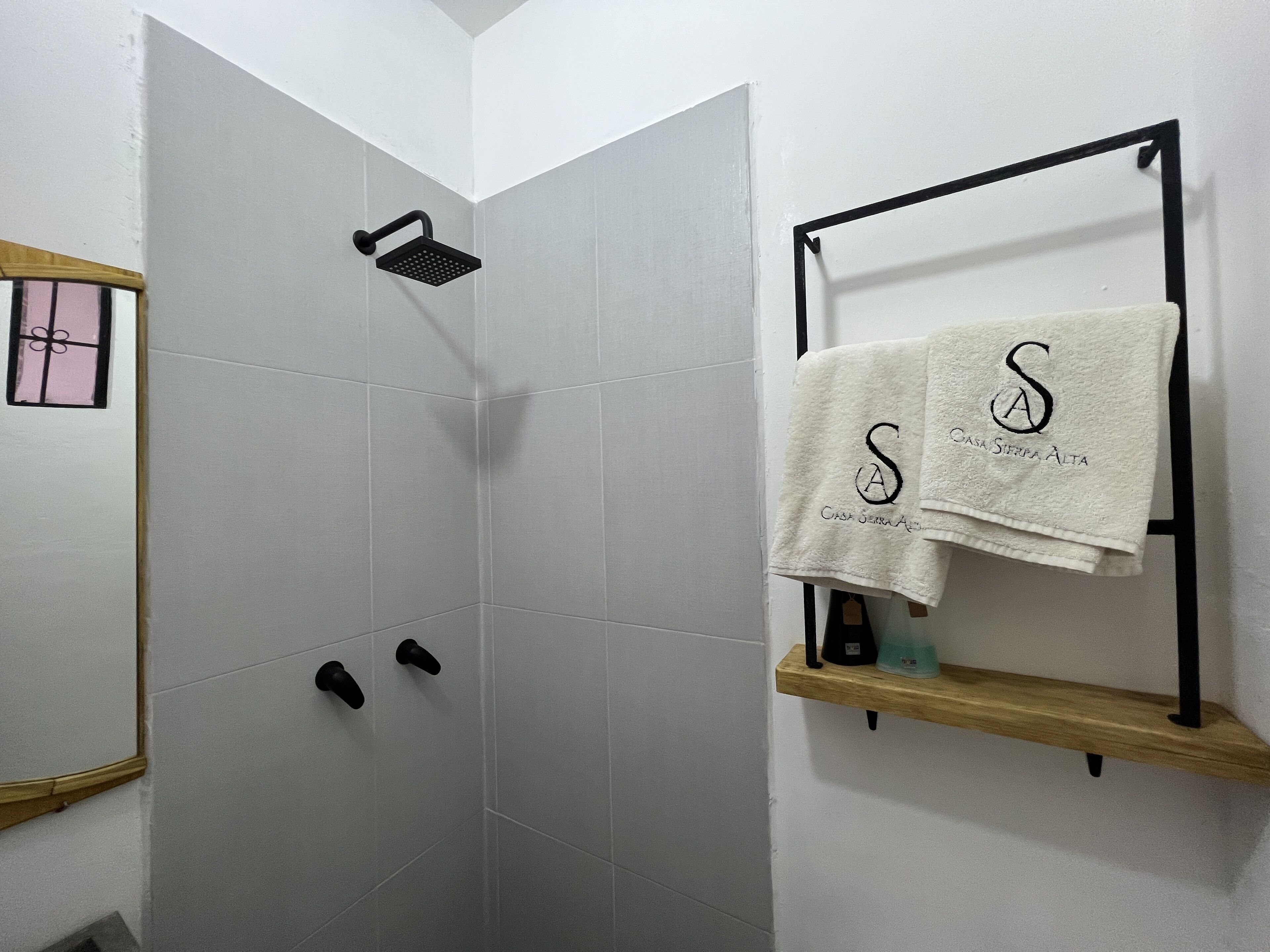 A shower area with a black handheld showerhead, a towel rack with two towels, and a wooden shelf with toiletries.