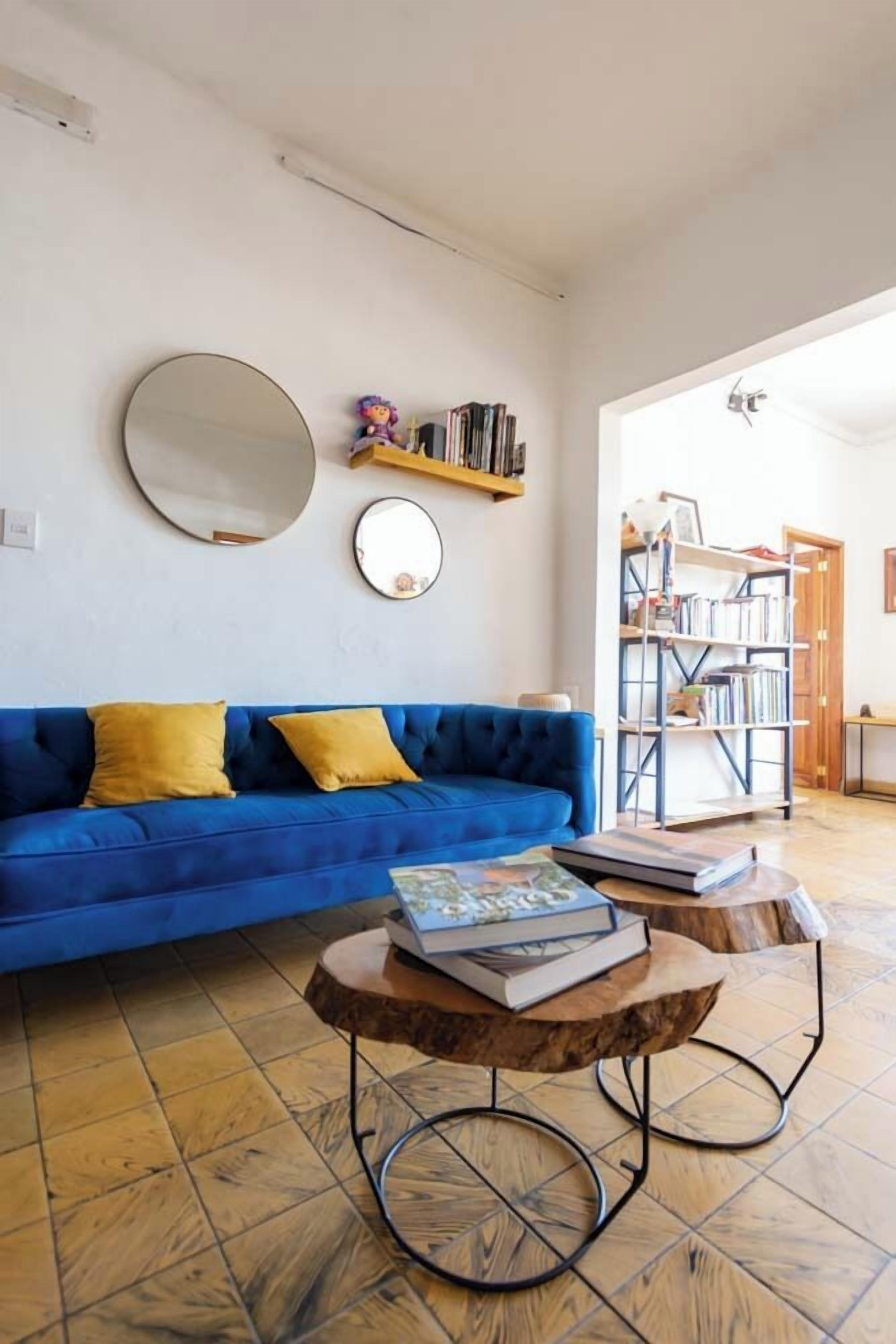 A living room with a blue sofa, yellow cushions, a round mirror, and a bookshelf.