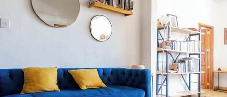 A living room with a blue sofa, yellow cushions, a round mirror, and a bookshelf.