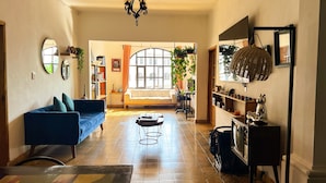 Amplia sala de estar con un sofá azul, una mesa de centro y un mueble para televisión. Hay un ventanal con cortinas, una estantería y una maceta colgante.