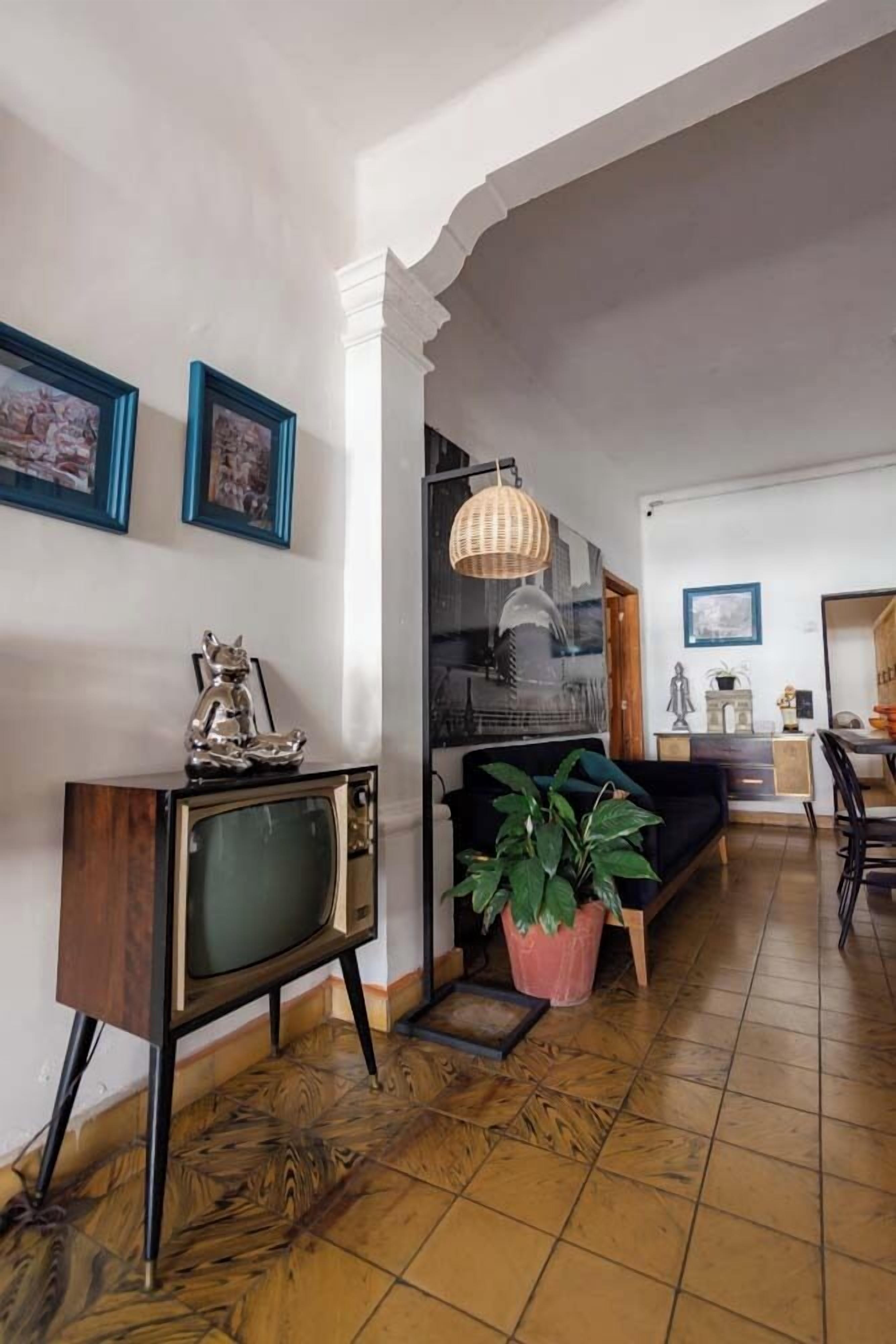 Una sala de estar con un televisor antiguo, una planta en maceta y un comedor con mesa y sillas.