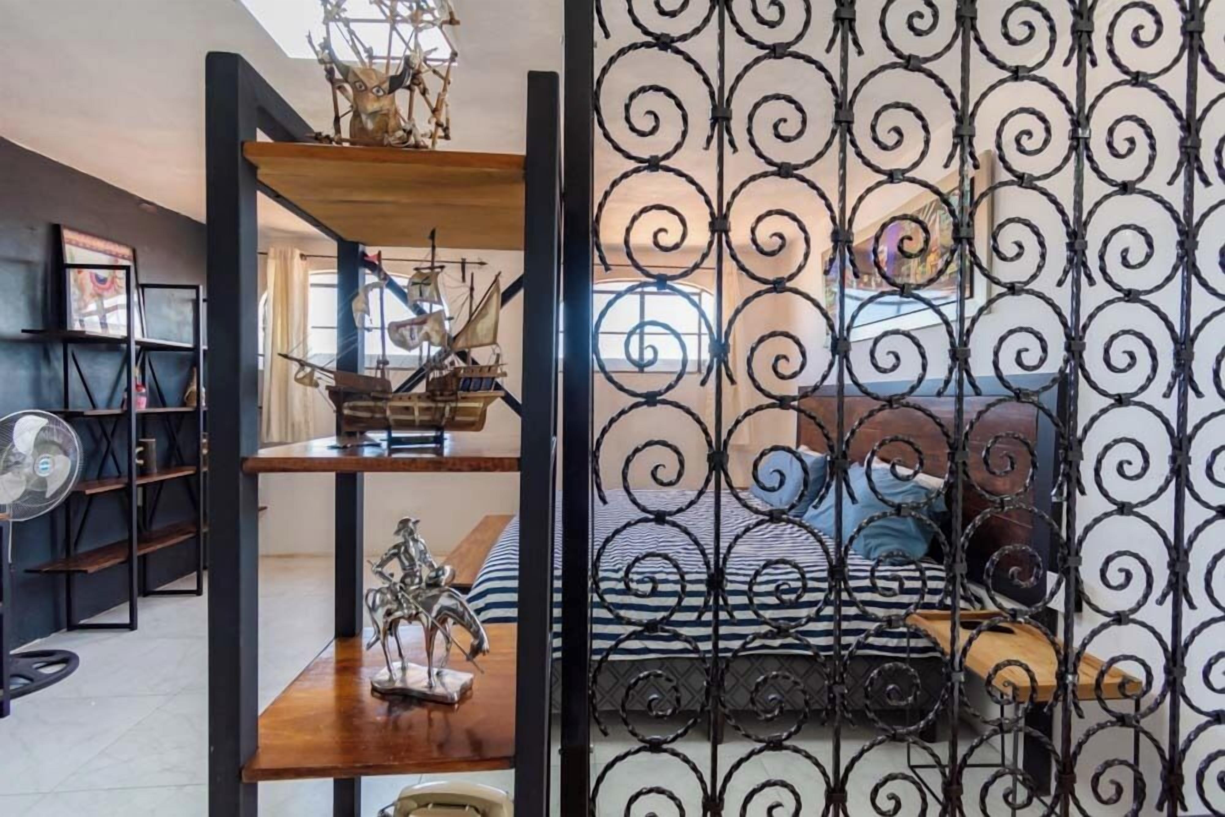 Quarto com cama, divisória metálica decorativa e estantes com diversos objetos, incluindo um modelo de navio e uma estátua.