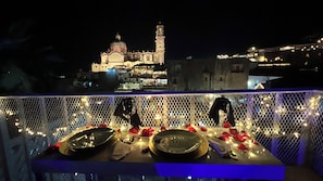 A dining table set with plates, cutlery, and a glass, overlooking a historic building with a bell tower at night.