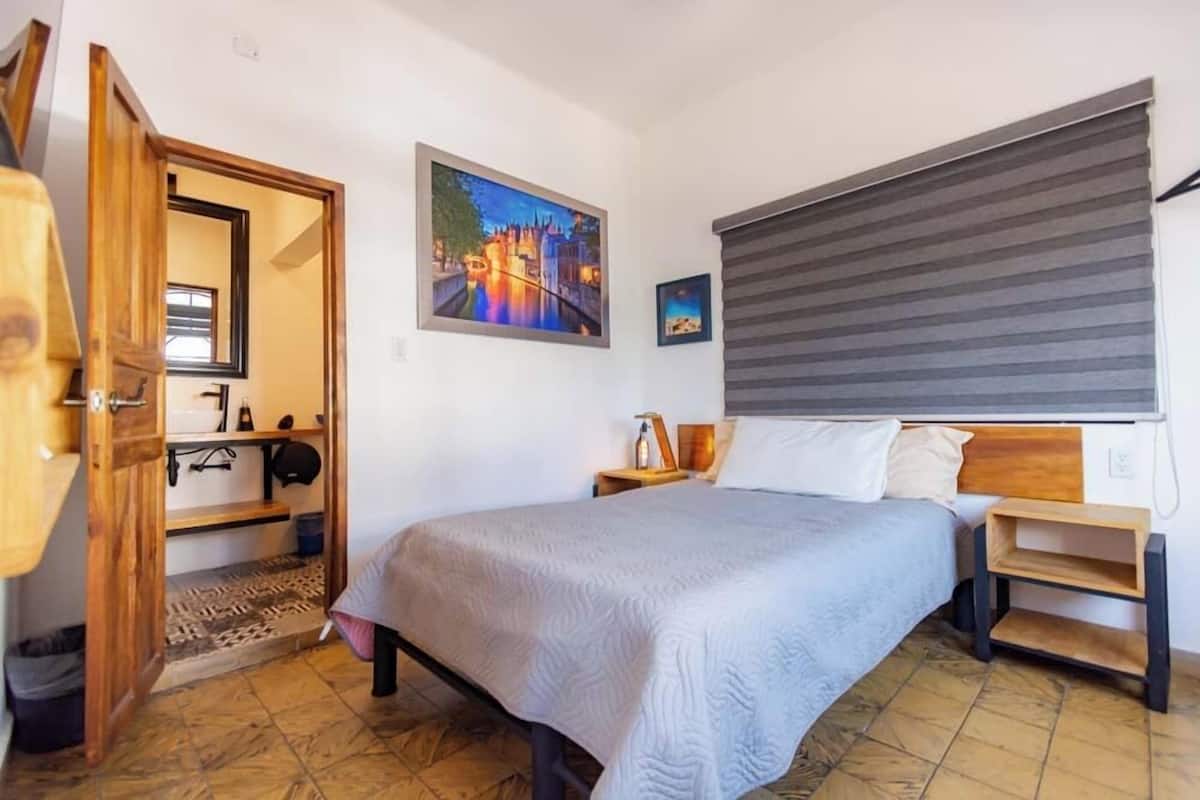 A bedroom with a bed, a wooden door, a painting, a window with blinds, and a tiled floor.
