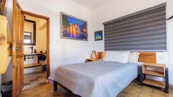 Ein Schlafzimmer mit einem Bett, einer Holztür, einem Gemälde, einem Fenster mit Jalousien und einem Fliesenboden.