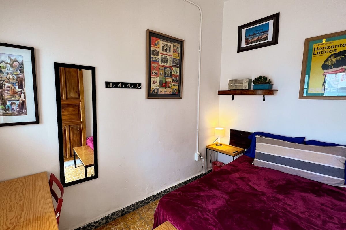 A bedroom with a bed, a desk, a chair, a mirror, and several framed pictures on the wall.