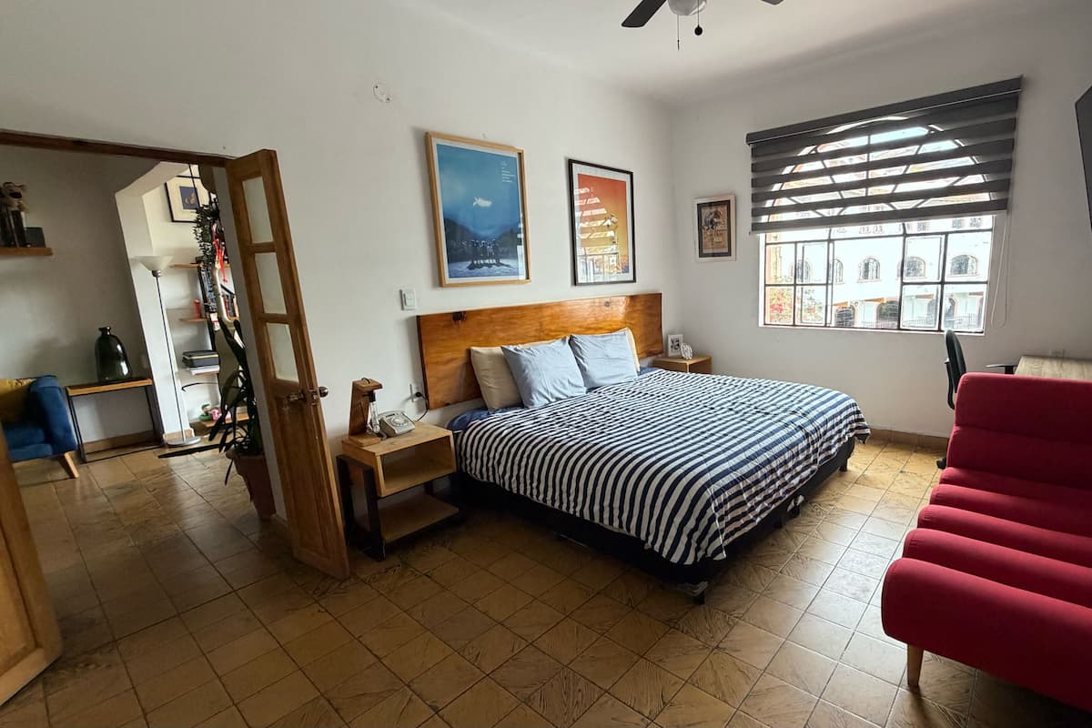 A bedroom with a bed, a nightstand, a red chair, a wooden headboard, a window with blinds, and a door leading to another room.