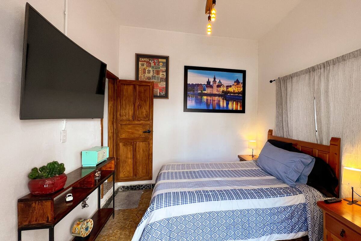A bedroom with a bed, a TV, a wooden wardrobe, a nightstand, a lamp, and a cityscape picture on the wall.