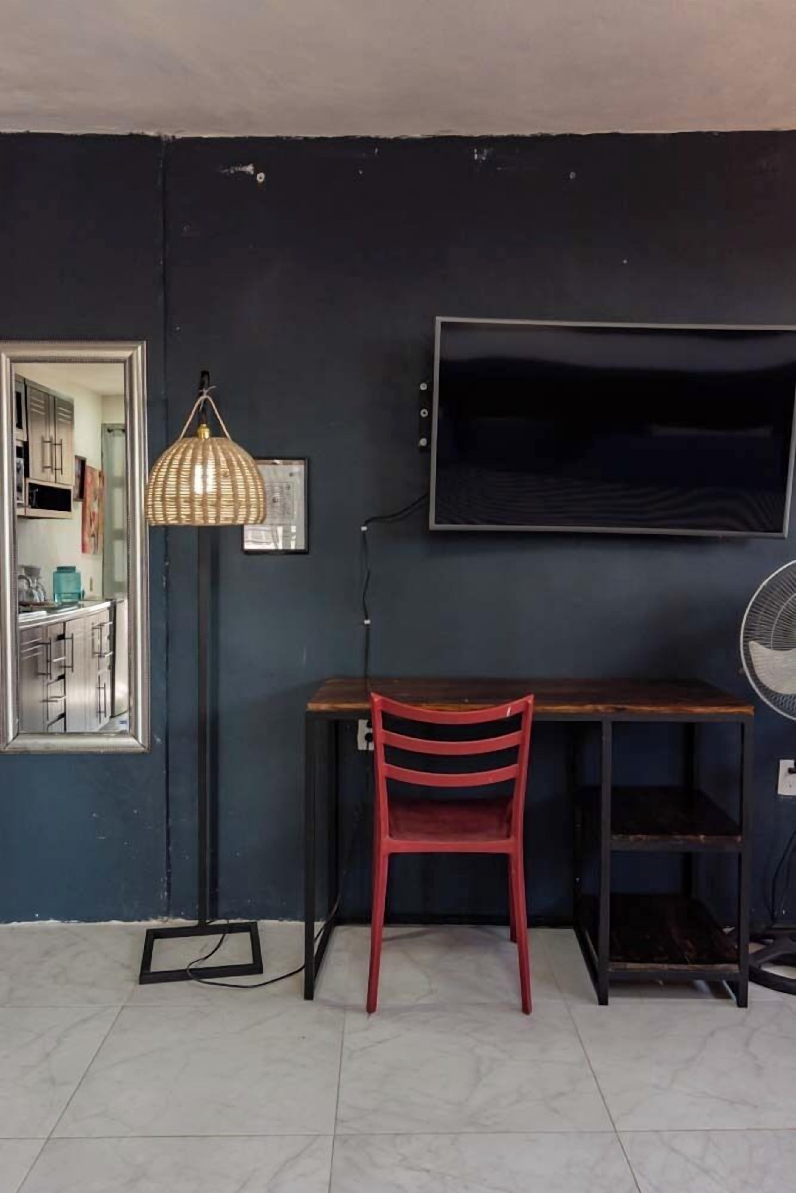 A room with a dark wall, a wooden desk, a red chair, a floor lamp, a TV, and a mirror reflecting a kitchen area.