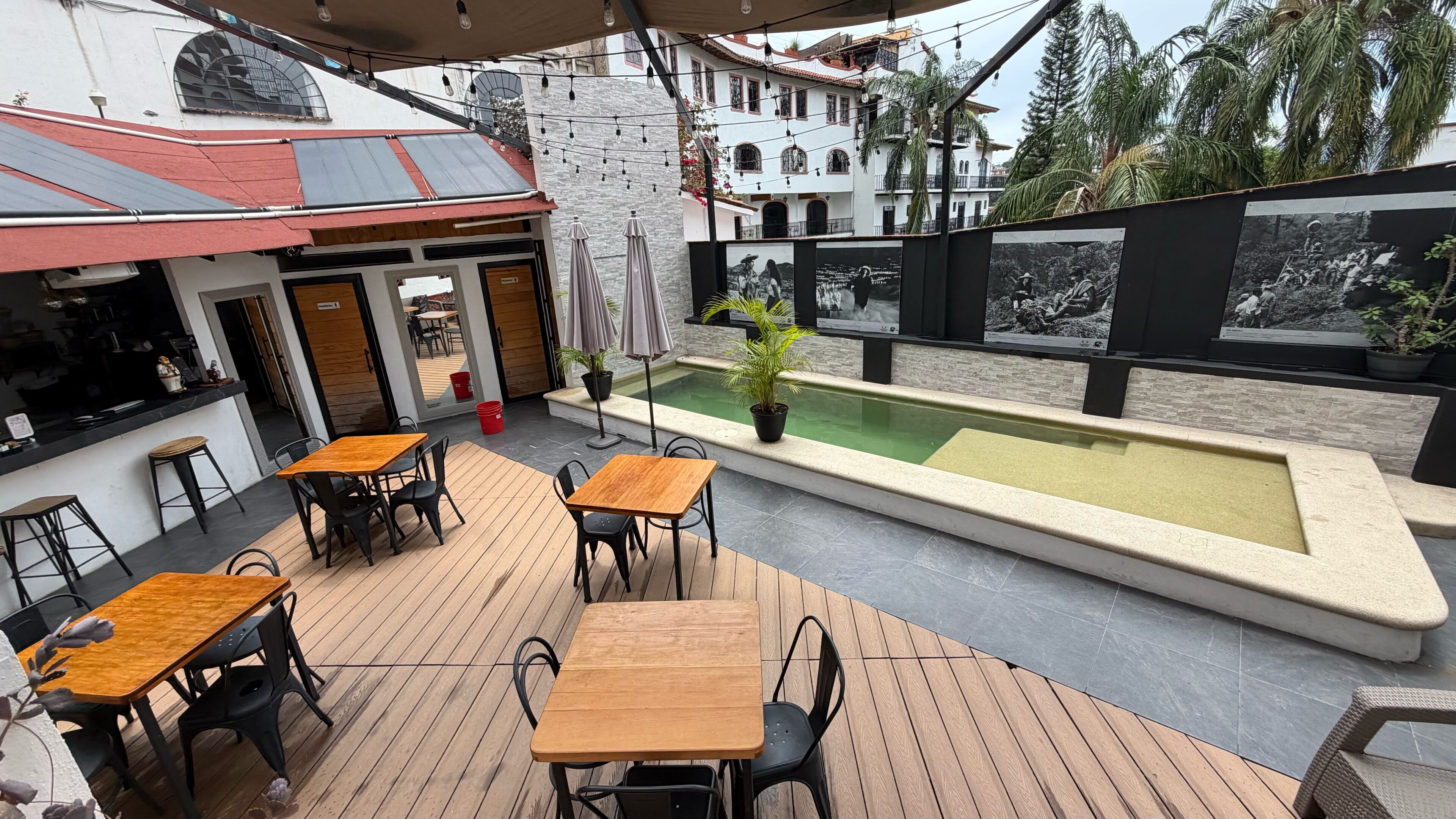Un patio al aire libre con mesas y sillas de madera, una piscina pequeña y vistas a un edificio tradicional con ventanas arqueadas.