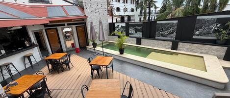 Un patio al aire libre con mesas y sillas de madera, una piscina pequeña y vistas a un edificio tradicional con ventanas arqueadas.