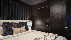 Un dormitorio con cama, cortinas oscuras, una puerta y una mesita de noche con lámpara.