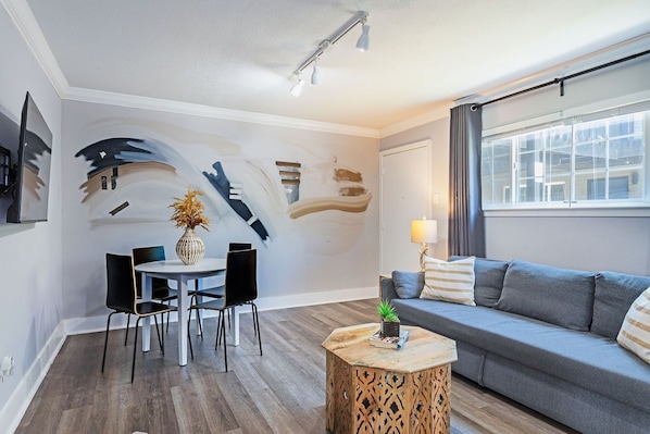 Una sala moderna con un sofá gris, una mesa de centro redonda de madera, una mesa de comedor con sillas y una pared decorada con arte abstracto.
