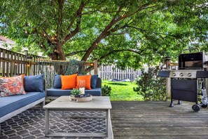Une terrasse extérieure avec une plateforme en bois, un canapé bleu avec des coussins orange, une table basse avec un plateau en osier et un barbecue.