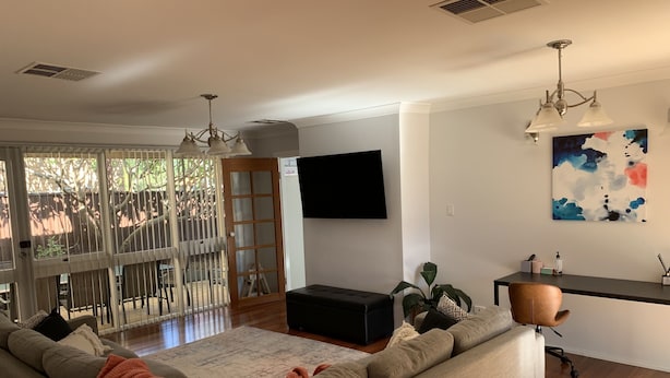 A modern living room with a grey sectional sofa, a coffee table, a TV mounted on the wall, and a large window with blinds.