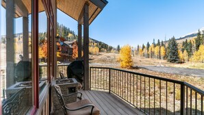 Una terraza de madera con mobiliario exterior, una parrilla y vistas a una zona boscosa con follaje otoñal.