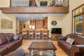 A spacious living room with leather sofas, a wooden coffee table, and a kitchen area with wooden cabinets and a television.