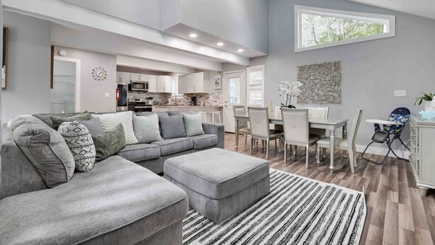 A spacious living room with a grey sectional sofa, a dining area with a white table and chairs, and a kitchen in the background. - Modern Poconos Central Cozy 5BR Home - Game Room (Tobyhanna)