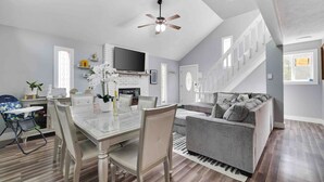A spacious living area with a dining table, a gray sofa, and a TV mounted on the wall. - Modern Poconos Central Cozy 5BR Home - Game Room (Tobyhanna)