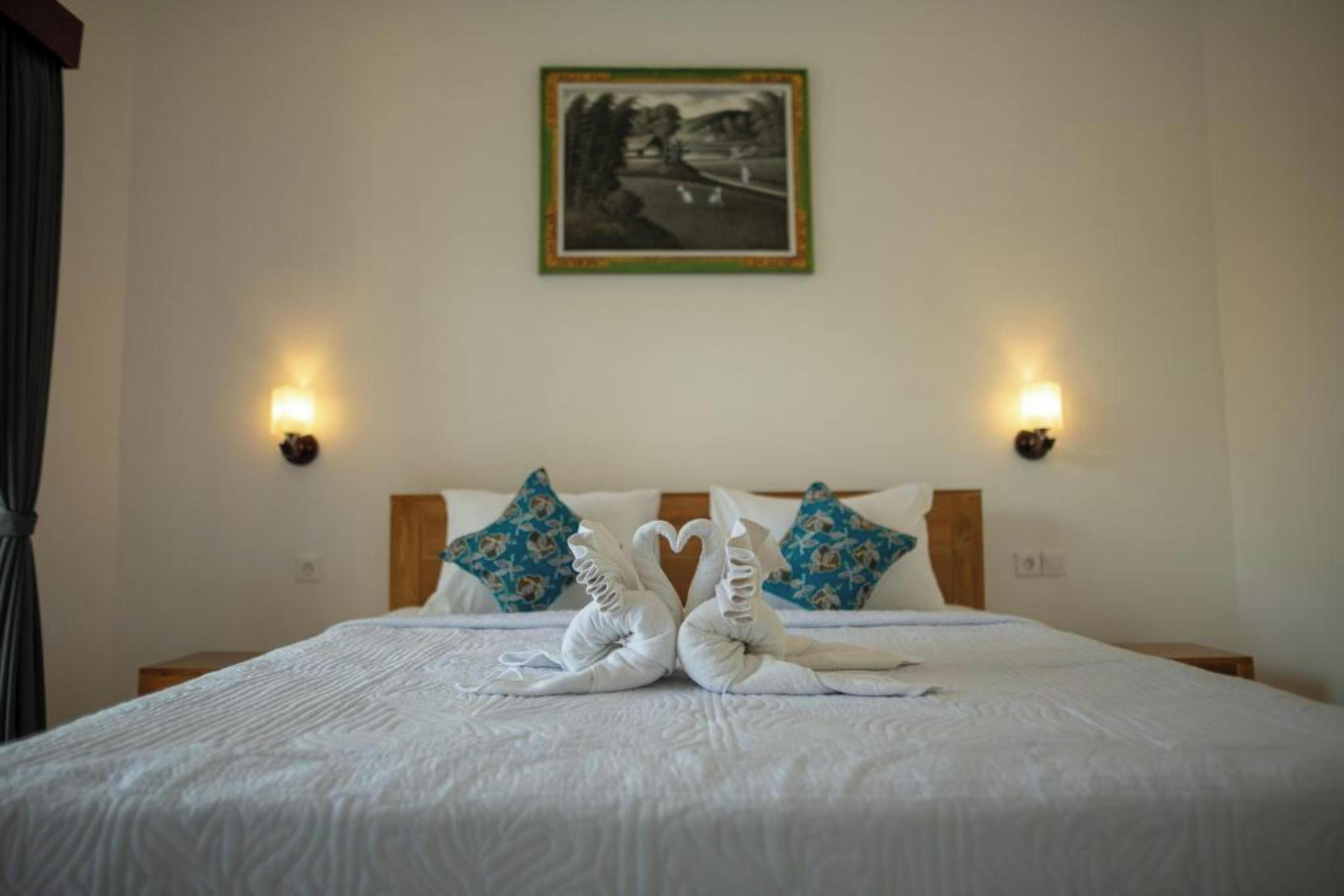 Cama arrumada com duas toalhas dispostas em formato de coração, um quadro na parede e duas luminárias fixadas na parede.