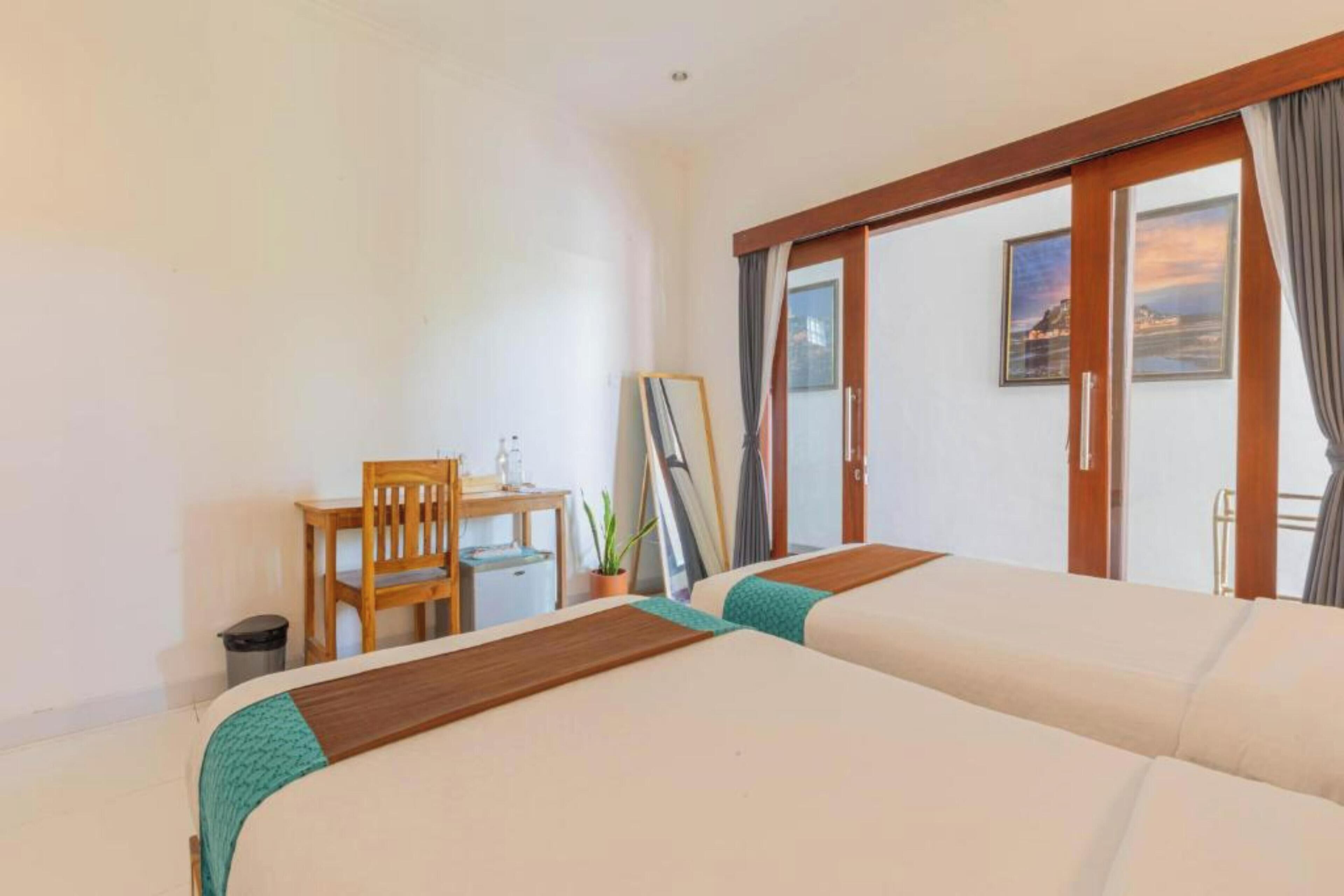A hotel room with two beds, a desk, a chair, a small table, a trash bin, and a sliding glass door leading to a balcony.
