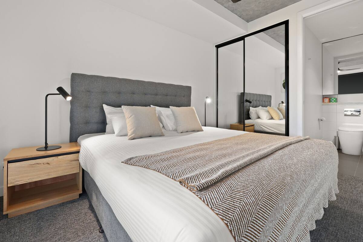 A modern bedroom with a large bed, a nightstand, a desk, and a mirror.