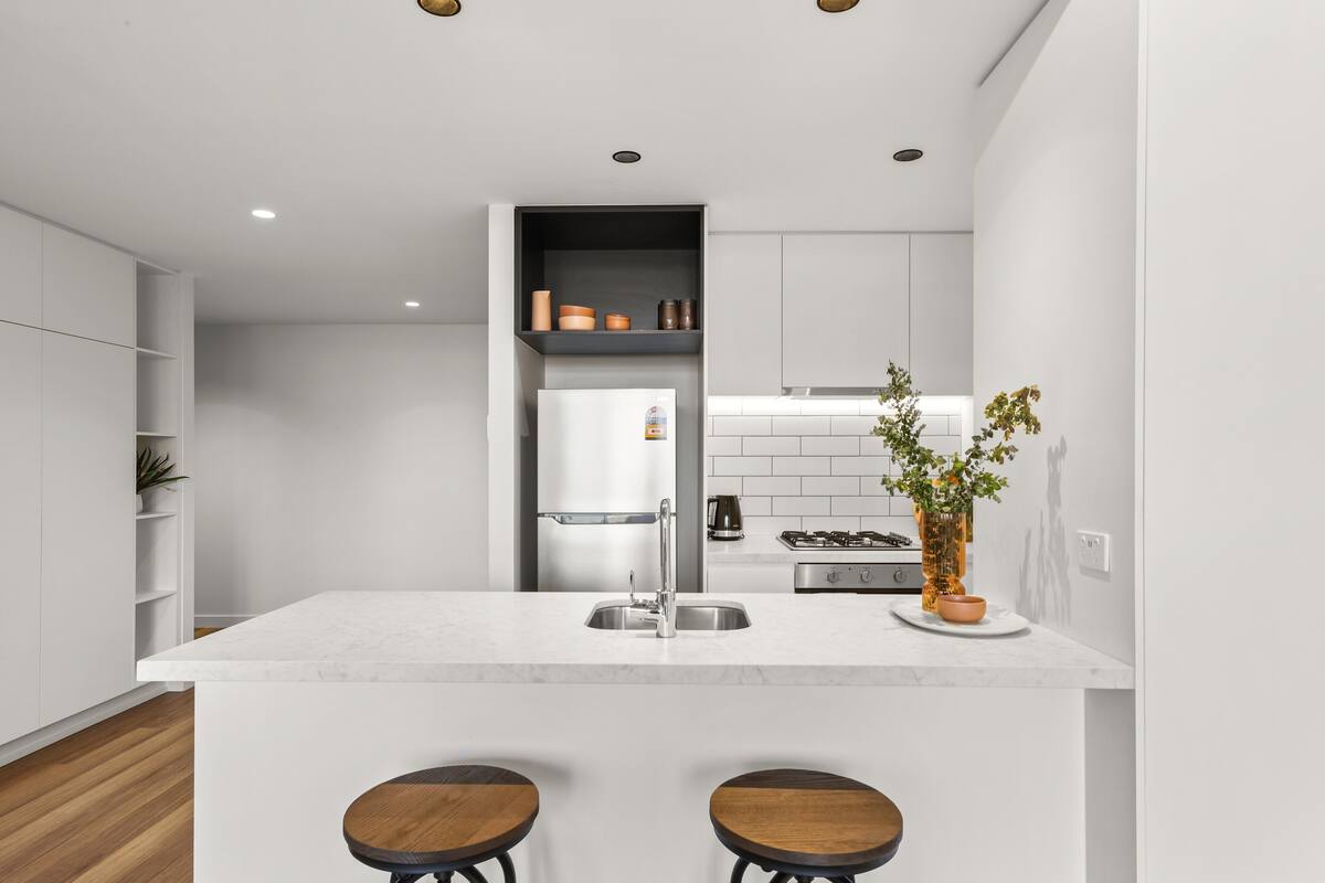 A modern kitchen with a central island, two stools, a built-in oven, and a potted plant.