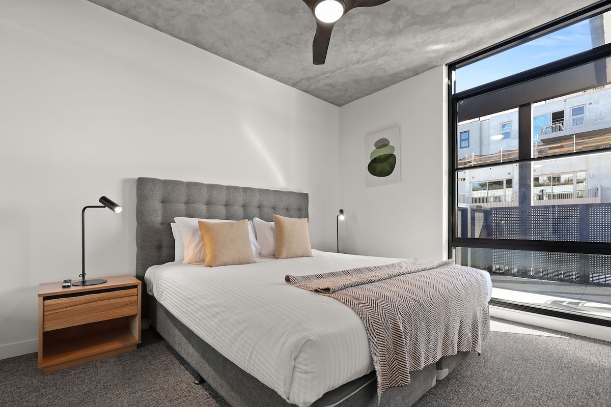 A modern bedroom with a large bed, two bedside tables, a ceiling fan, and a view of the city through large windows.