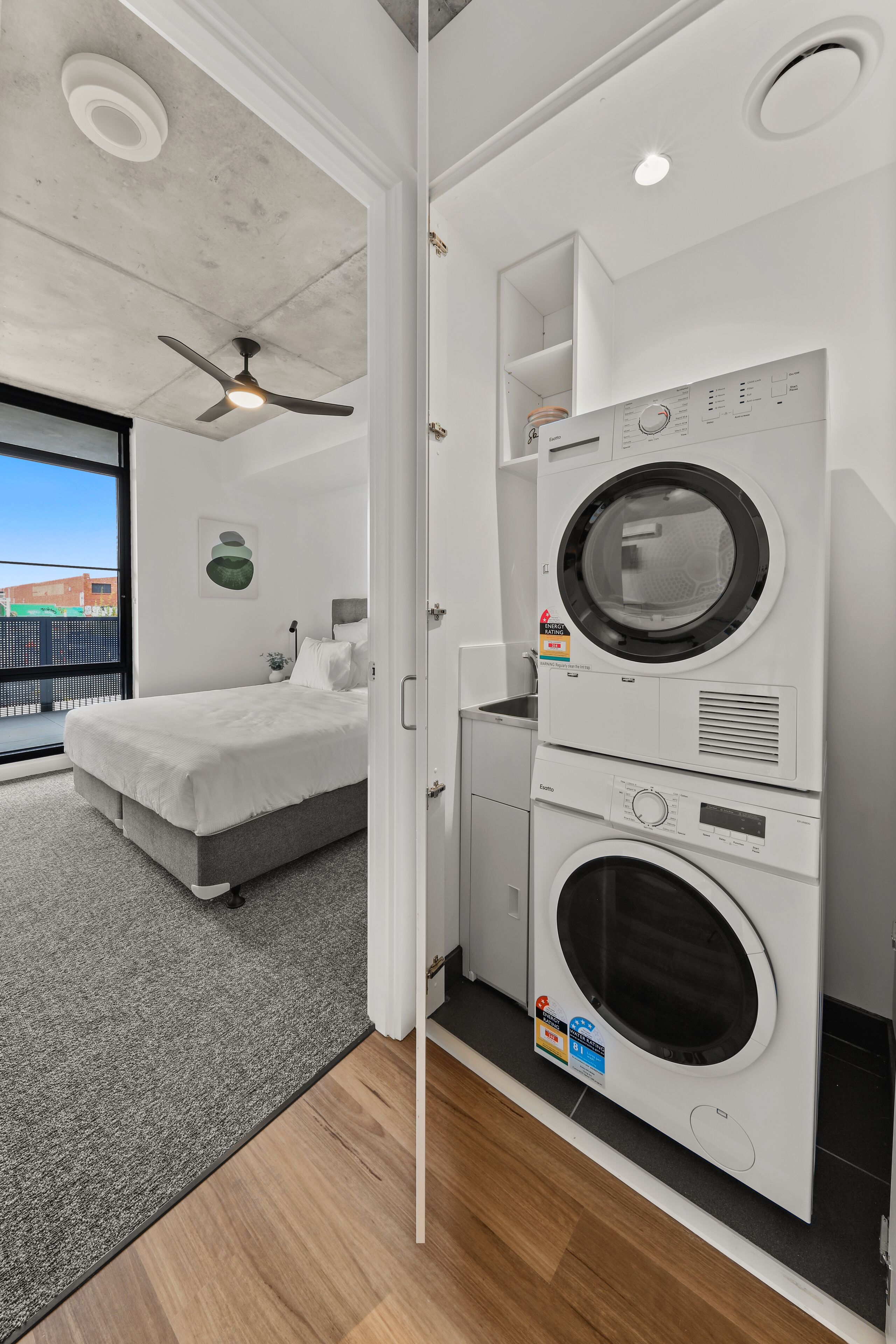 Une petite buanderie équipée d’un lave-linge et d’un sèche-linge encastrés, d’un lit gris et d’un ventilateur de plafond.
