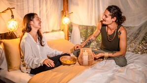 A couple sitting on a bed in a tented area with a woven basket and a small drum between them. - Awaken Yoga Eco Resort in Amazon jungle (Indiana)