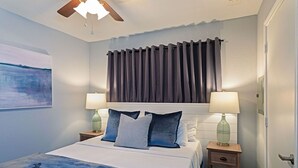 Un dormitorio con cama, dos mesitas de noche con lámparas, ventilador de techo y ventana con cortinas.