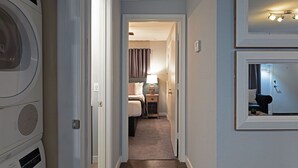 Un couloir étroit mène à une chambre où se trouvent un lit, une table de chevet et une lampe. On peut apercevoir un lave-linge sur la gauche.