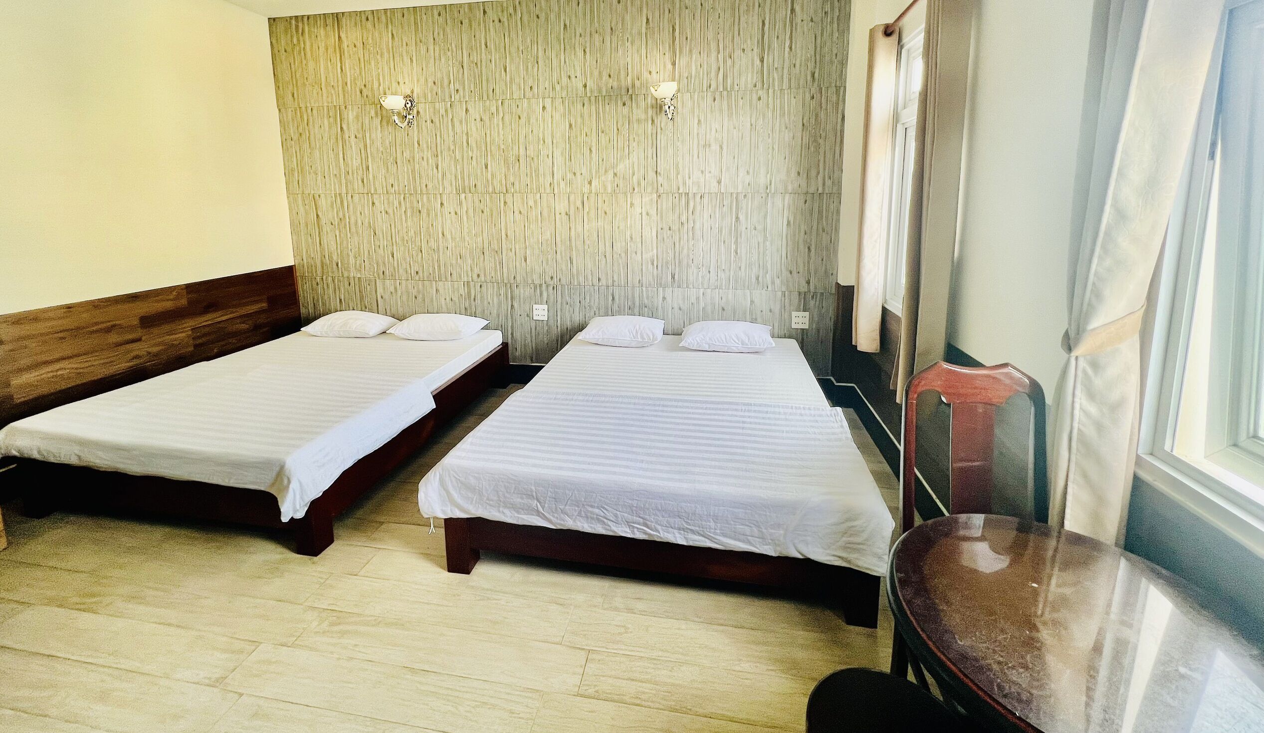 Ein Hotelzimmer mit zwei Einzelbetten, einem Holzkopfteil, hellen Wänden und einem Fenster mit Vorhängen.