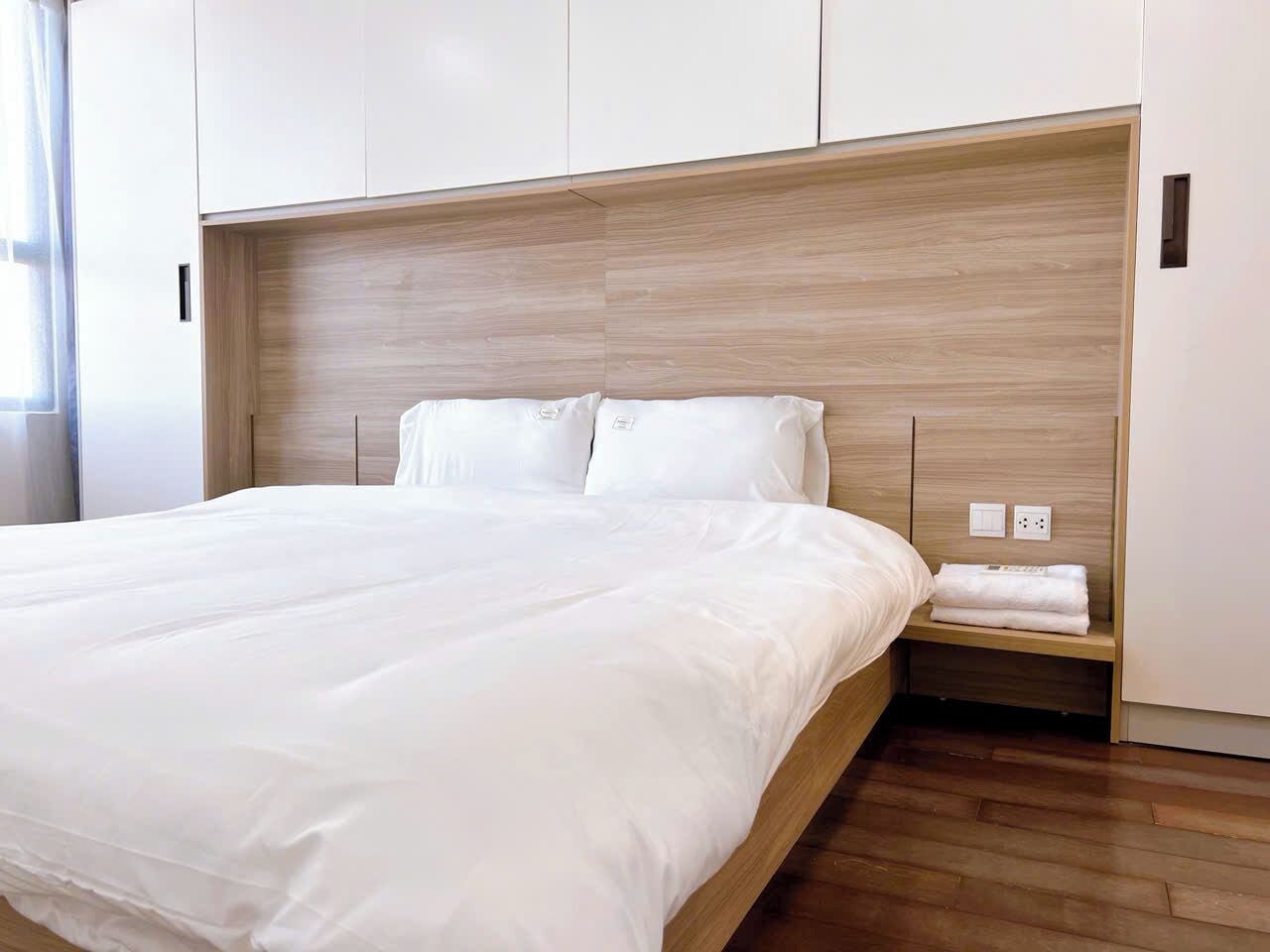 A neatly made bed with white linens in a modern room with wooden flooring and built-in wardrobes.