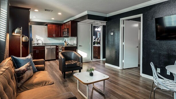 A modern living room with a brown leather sofa, a small white coffee table, and a black leather chair. There is a kitchen area with wooden cabinets and a microwave.