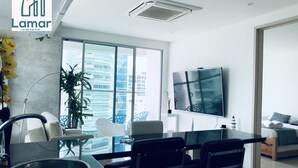 A modern hotel room with a large window, a flat-screen TV, a dining area with a glass table, and a bed with a view of the city. - Murano Elite Piso 25 (Cartagena de Indias)