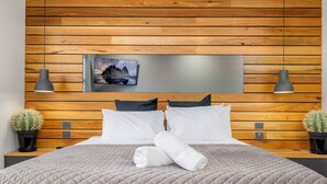 A neatly made bed with a quilted cover, two pillows, and a rolled towel. There are two bedside tables with lamps and potted cacti. A framed picture hangs on the wooden wall above the bed. - Jindy Snowy Mountains Retreat (Jindabyne)