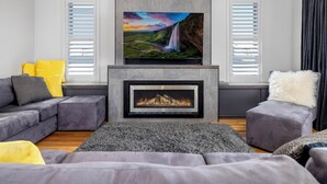 A modern living room with a fireplace, a large flat-screen TV mounted above it, and a cozy seating area with a gray sofa and ottoman. - Jindy Snowy Mountains Retreat (Jindabyne)
