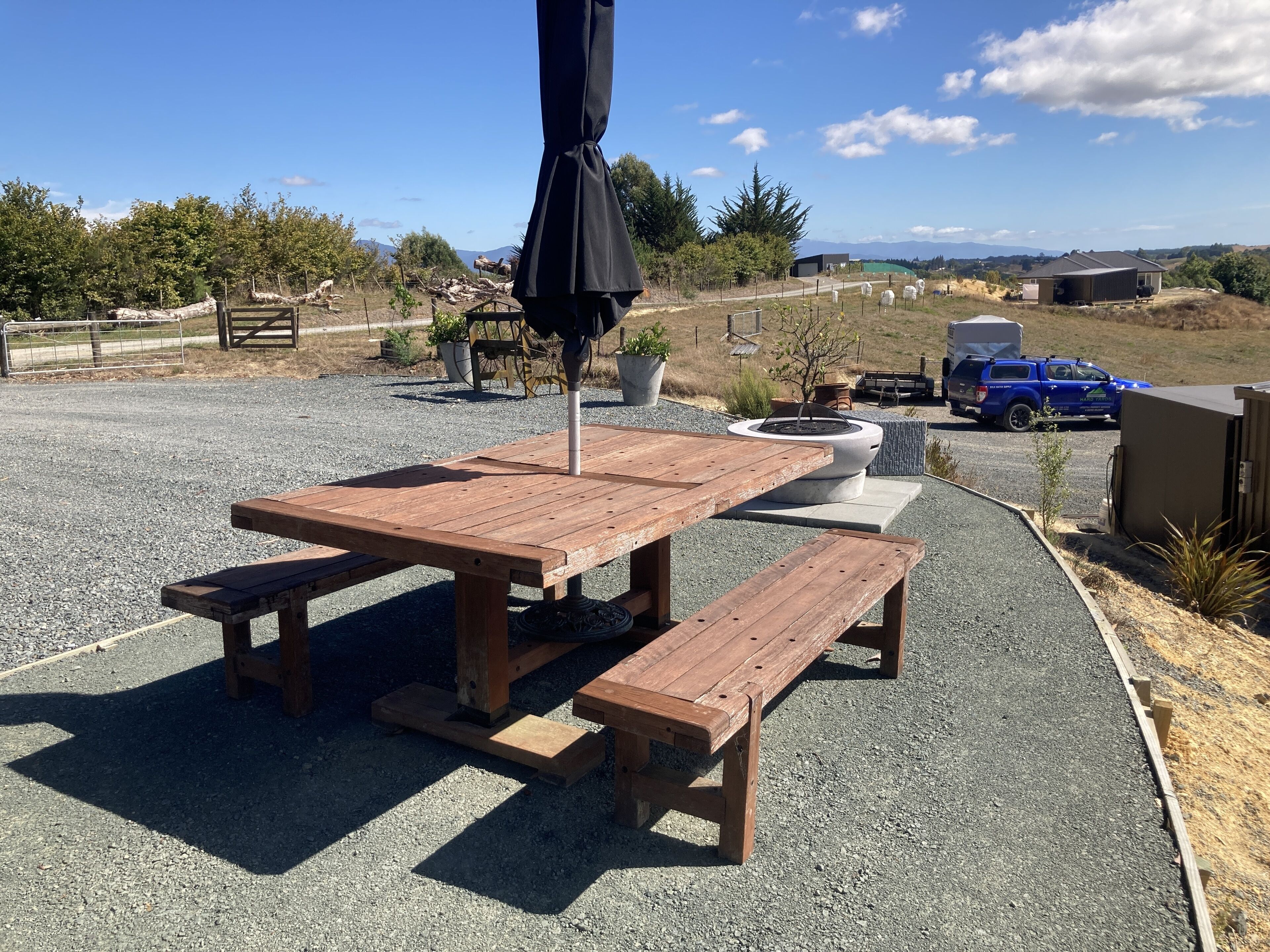 A wooden picnic table with benches, a fire pit, and a black umbrella.