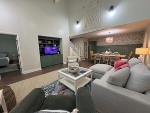 Uma sala de estar com televisão, sofá, mesa de centro, área de refeições com cadeiras e uma escada.