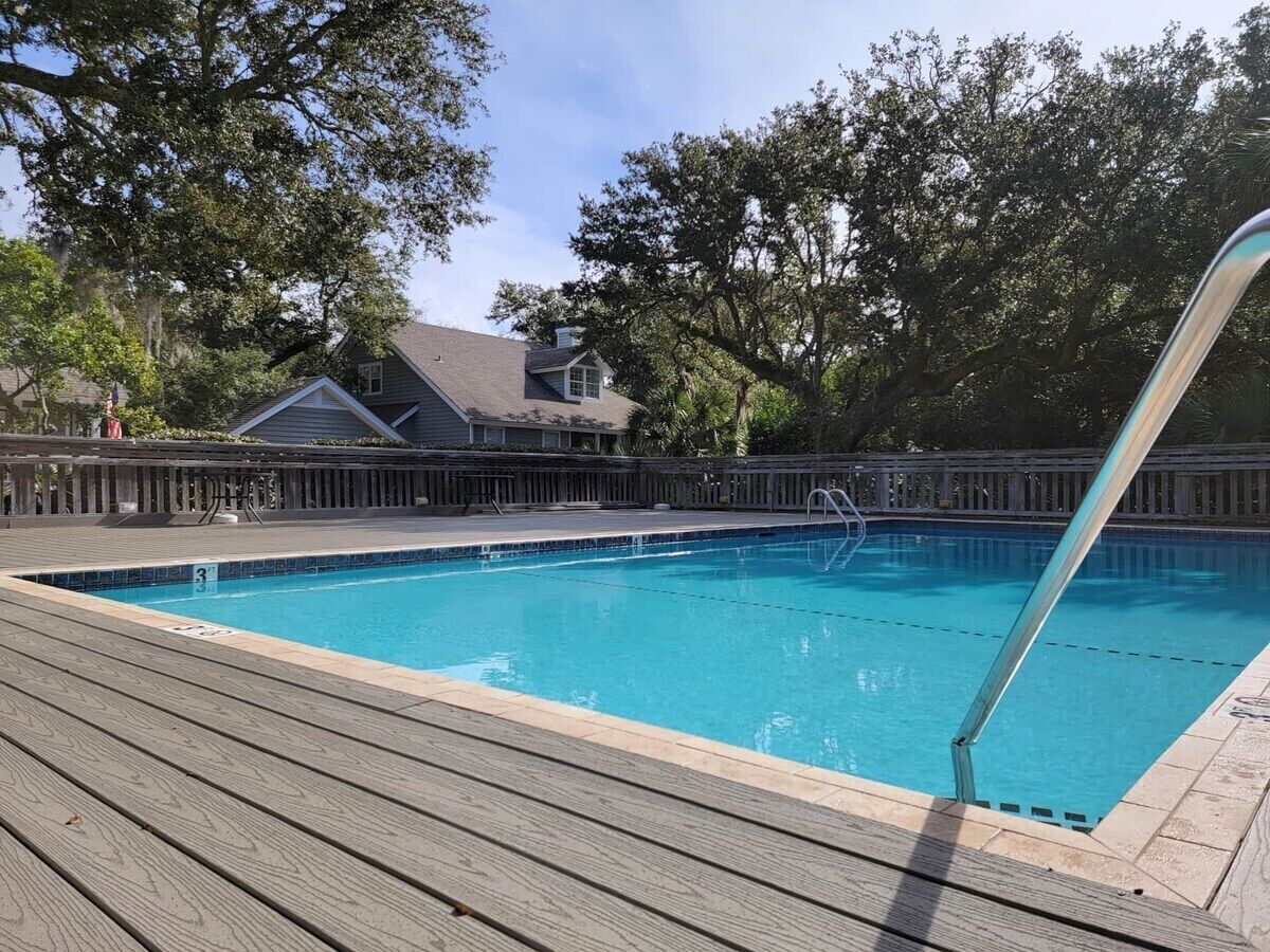 Une piscine avec une terrasse en bois, entourée d’arbres et avec une maison en arrière-plan.