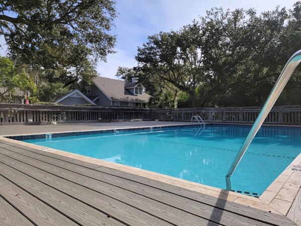 A swimming pool with a wooden deck, surrounded by trees and a house in the background.