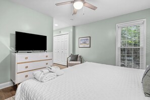 Un dormitorio con cama, cómoda, televisor, ventilador de techo, puerta, un cuadro enmarcado y una ventana con persianas.