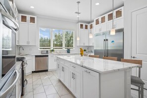 Une cuisine moderne avec des meubles blancs, un grand îlot central, des appareils électroménagers en acier inoxydable et une vue sur les arbres par les fenêtres.