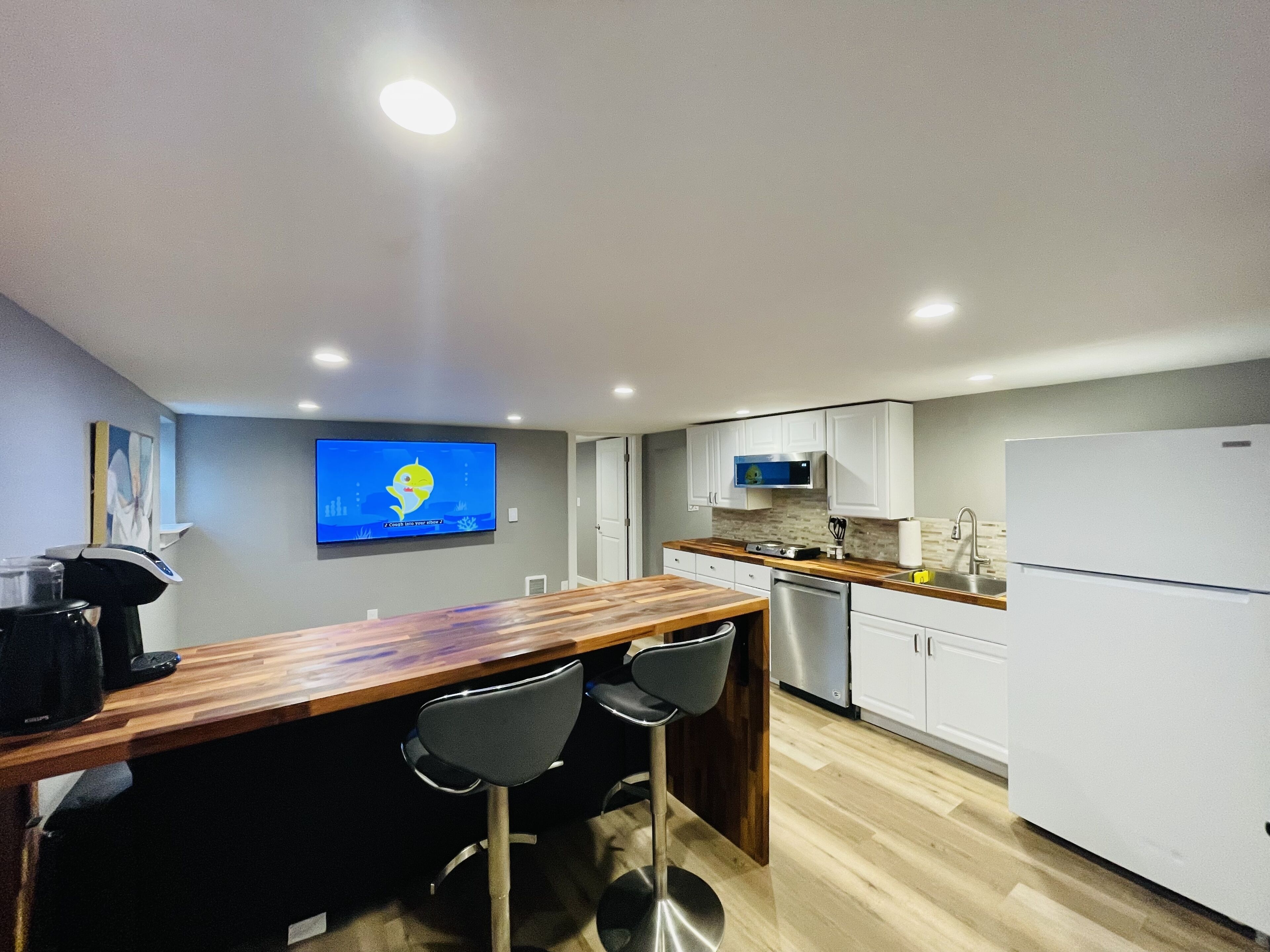 Une cuisine moderne avec un comptoir-bar en bois, deux tabourets de bar, des armoires blanches, un réfrigérateur en acier inoxydable et un téléviseur à écran plat fixé au mur.