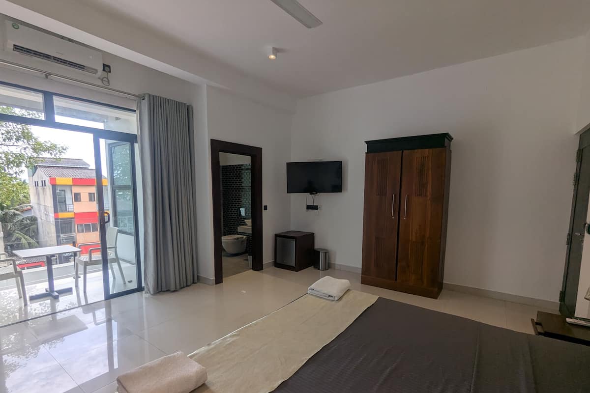 A modern hotel room with a large bed, a flat-screen TV, a wooden wardrobe, and a balcony with a table and chairs.
