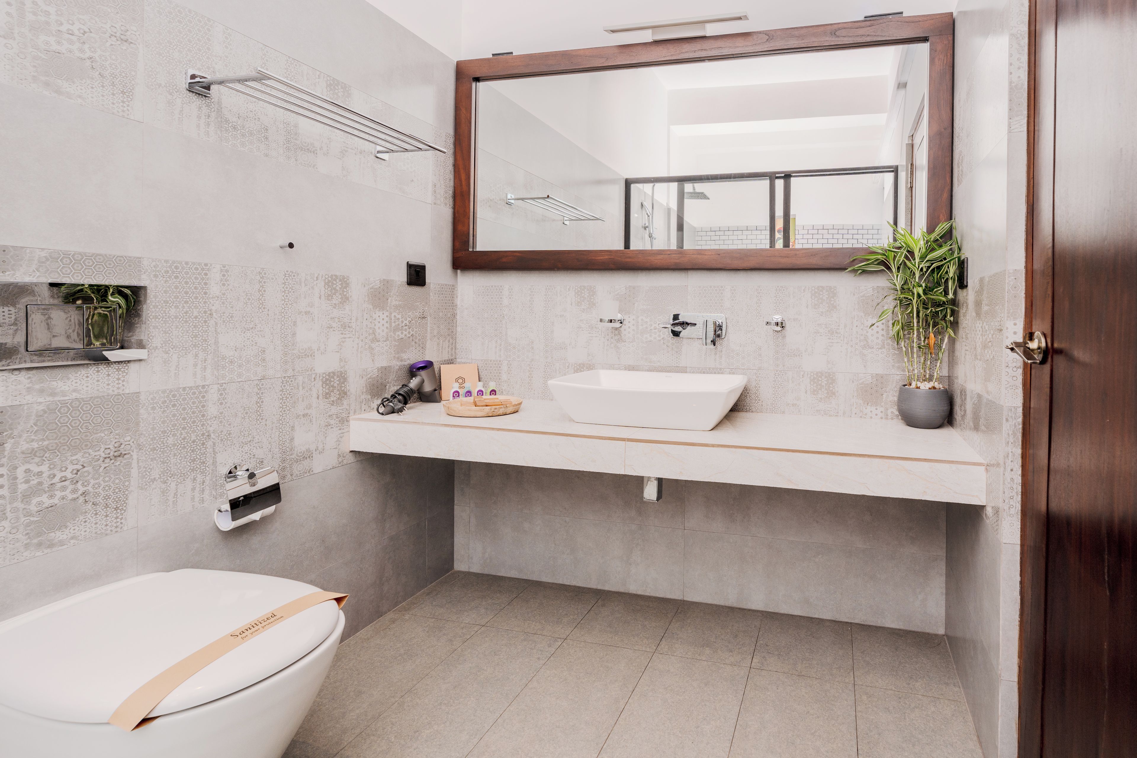 A modern bathroom with a white toilet, a wooden-framed mirror, a white sink, and a potted plant.