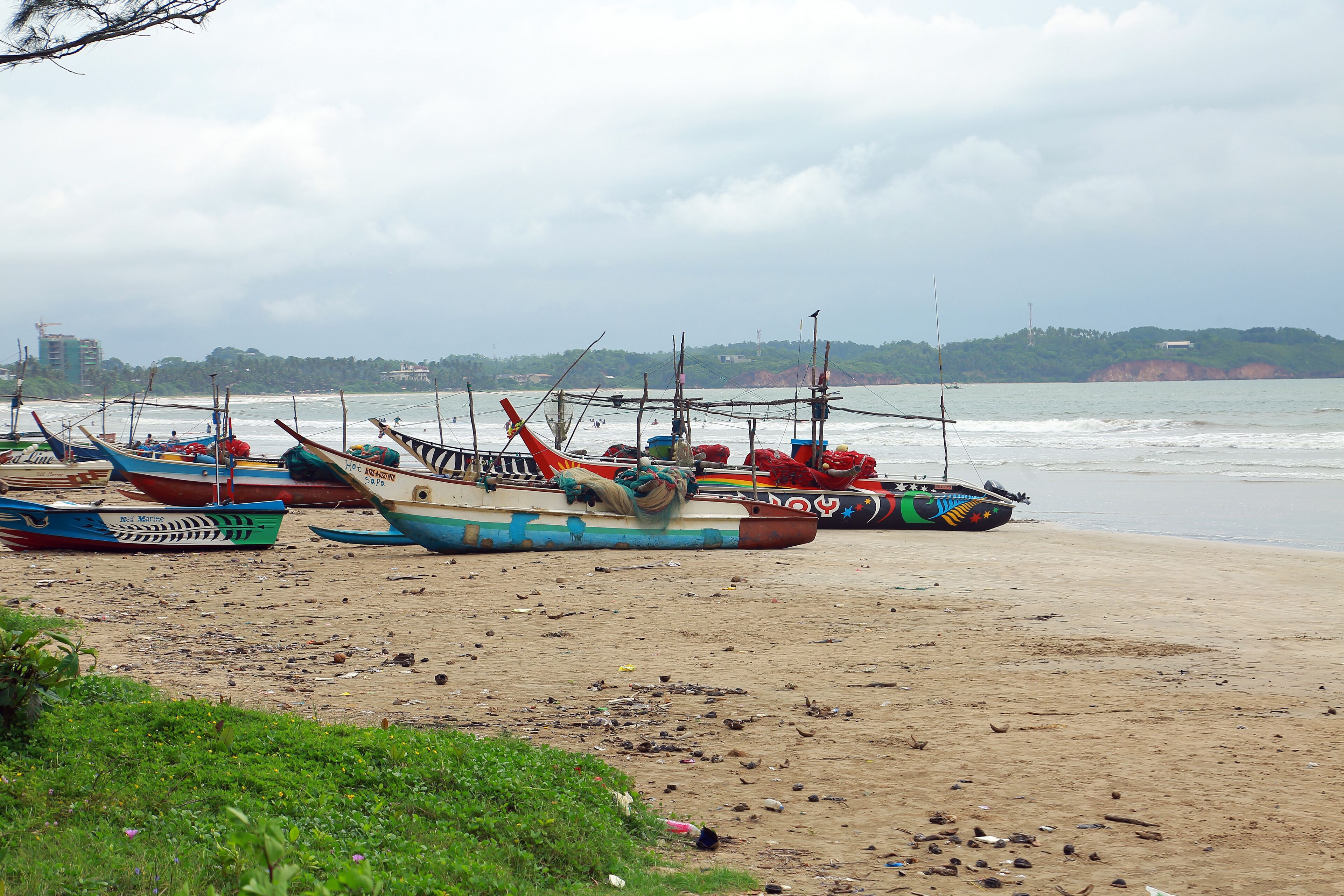 A beach with several boats moored on the sand, a distant shoreline with buildings, and a cloudy sky.