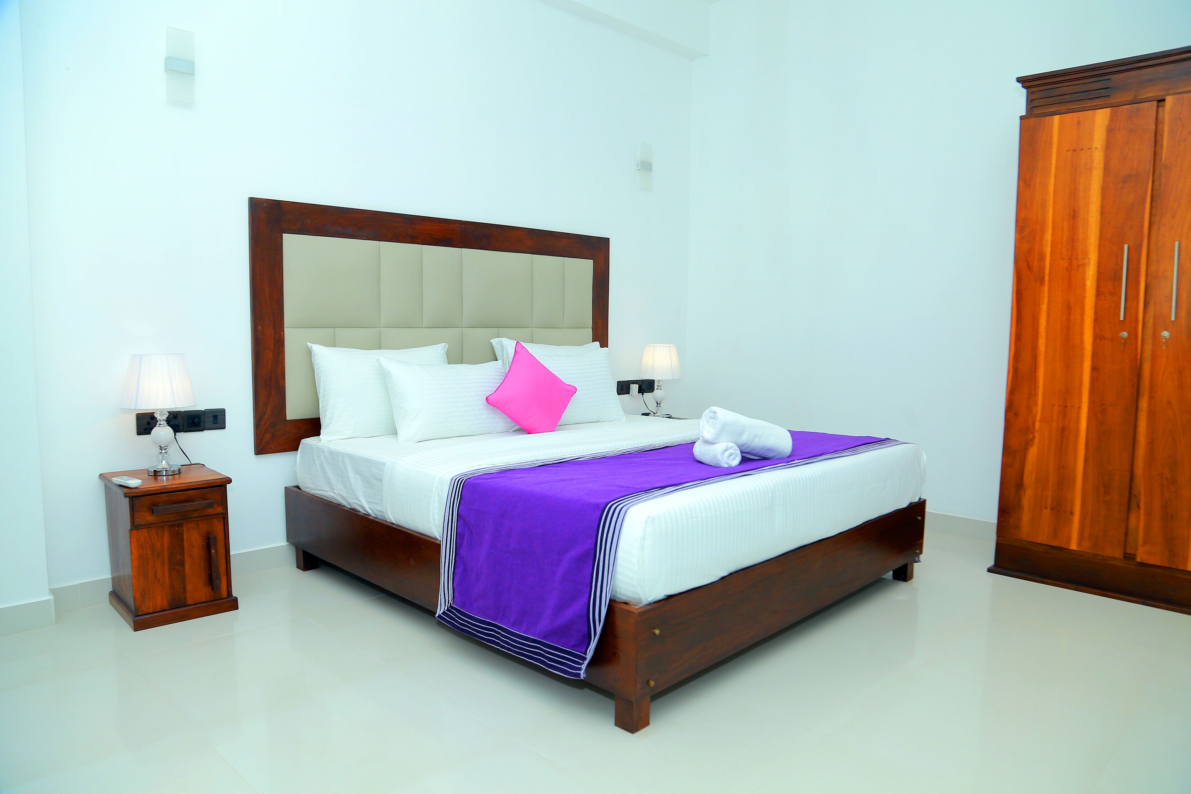 A neatly made bed with a wooden headboard, white bedding, a purple throw, and pink and white pillows. There are two bedside tables with lamps, and a wooden wardrobe to the right.